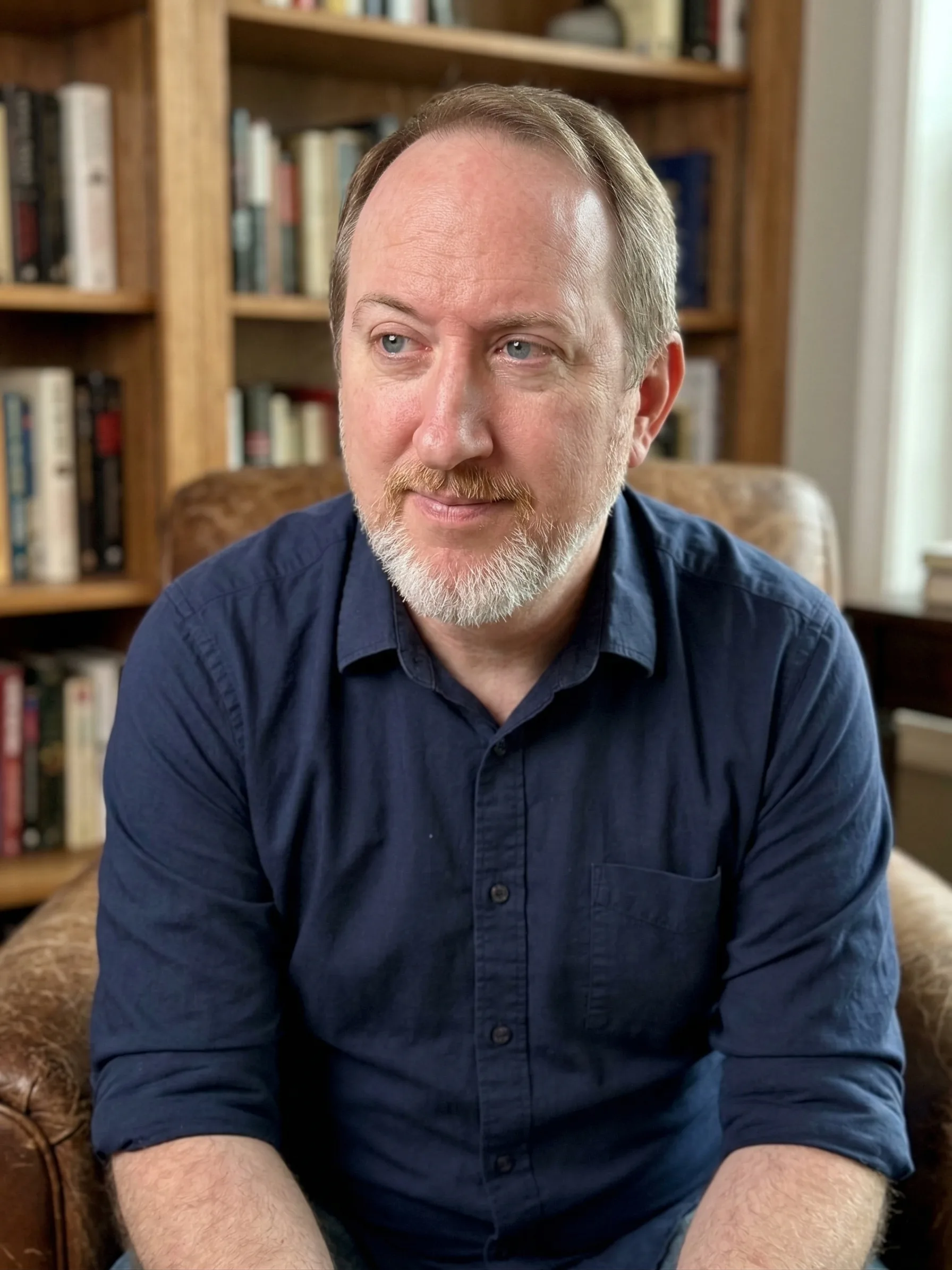 Philip Jarrell seated in a leather chair, in mid-thought, with a wall of books behind him